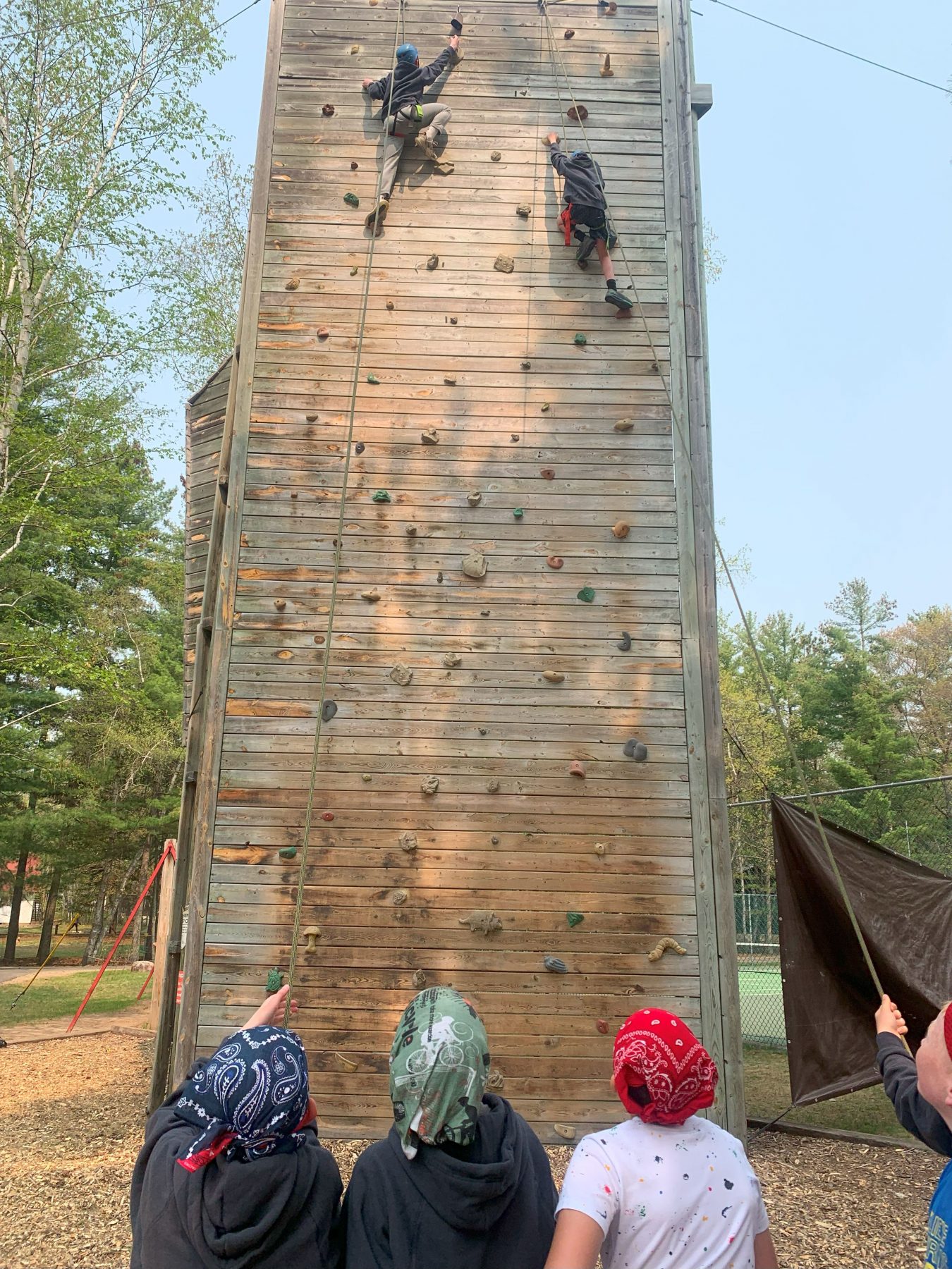 3 campers face away from the camera looking at a climbing wall with two climbers climbing up to the top 