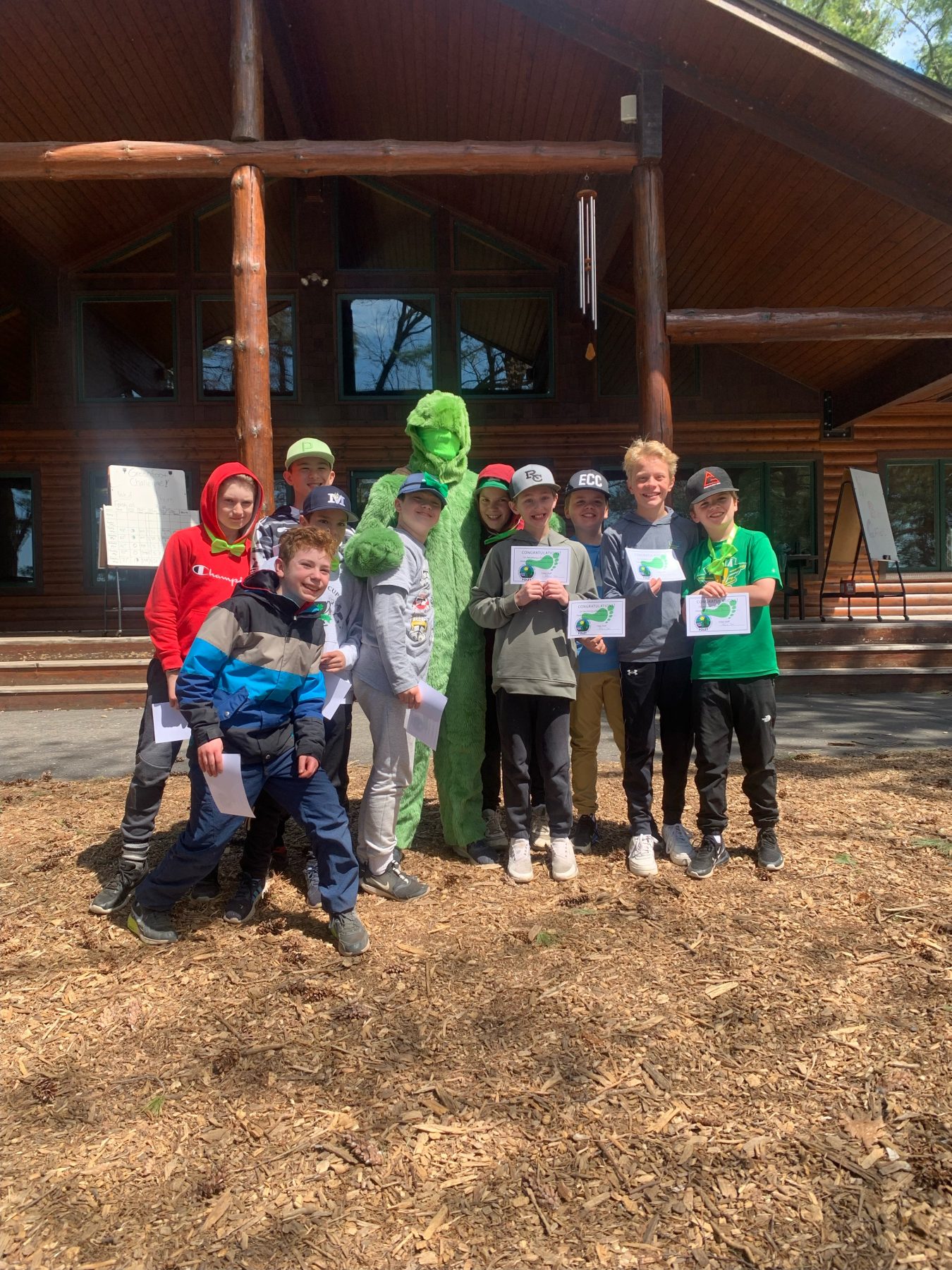 Kids stand around a camp counselor dressed as a fuzzy green monster, kids are smiling and holding certificates 