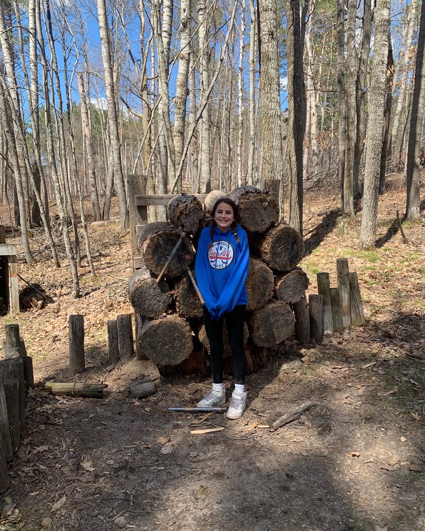 A girl in a blue sweatshirt stands in front of an axe throwing target with her axes stuck in 