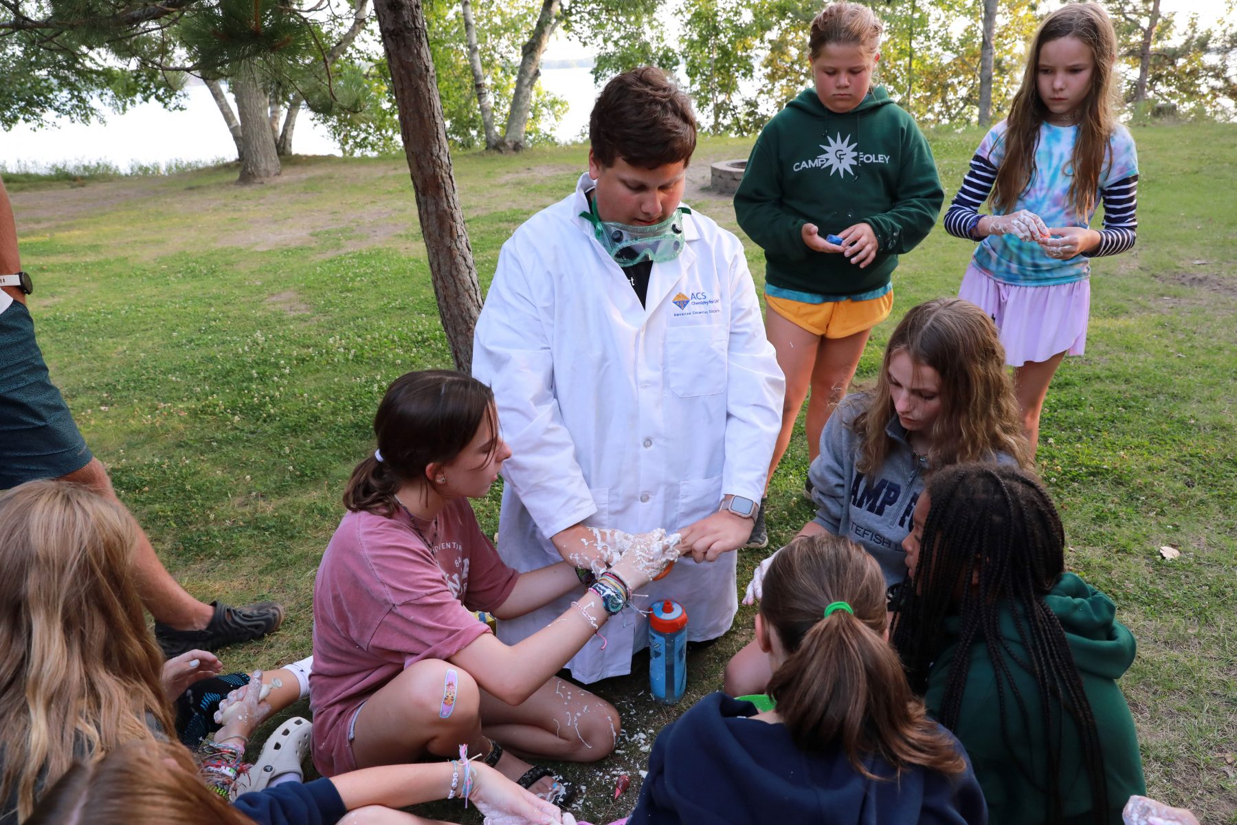 Man in a science lab coat sits with campers in a circle, with a girl to his right shows him her hands covered in slime at Camp Foley 