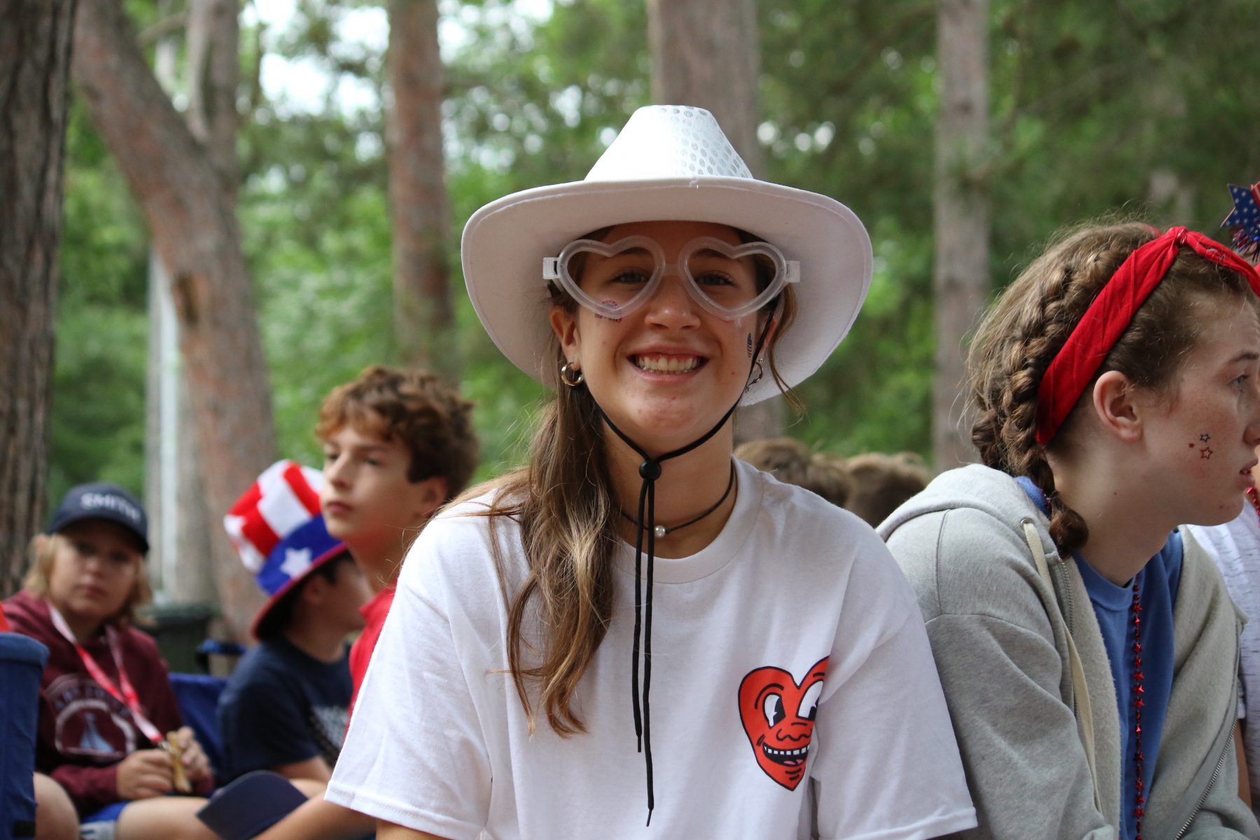 A young woman wears see through heart sunglasses and a cowboy hat, smiling at Camp Foley 