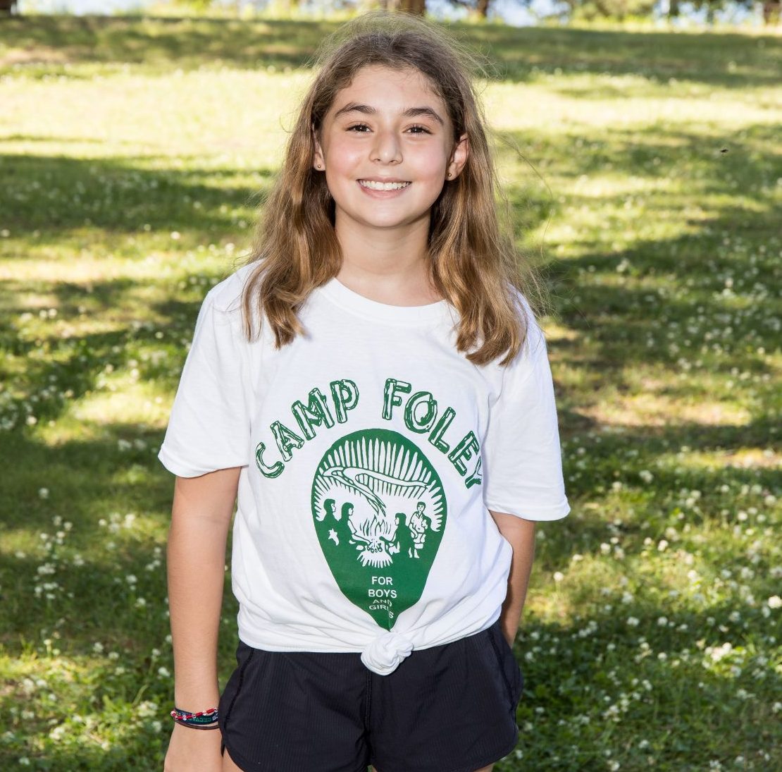 A young girl wear a white camp foley shirt while smiling and standing on grass 