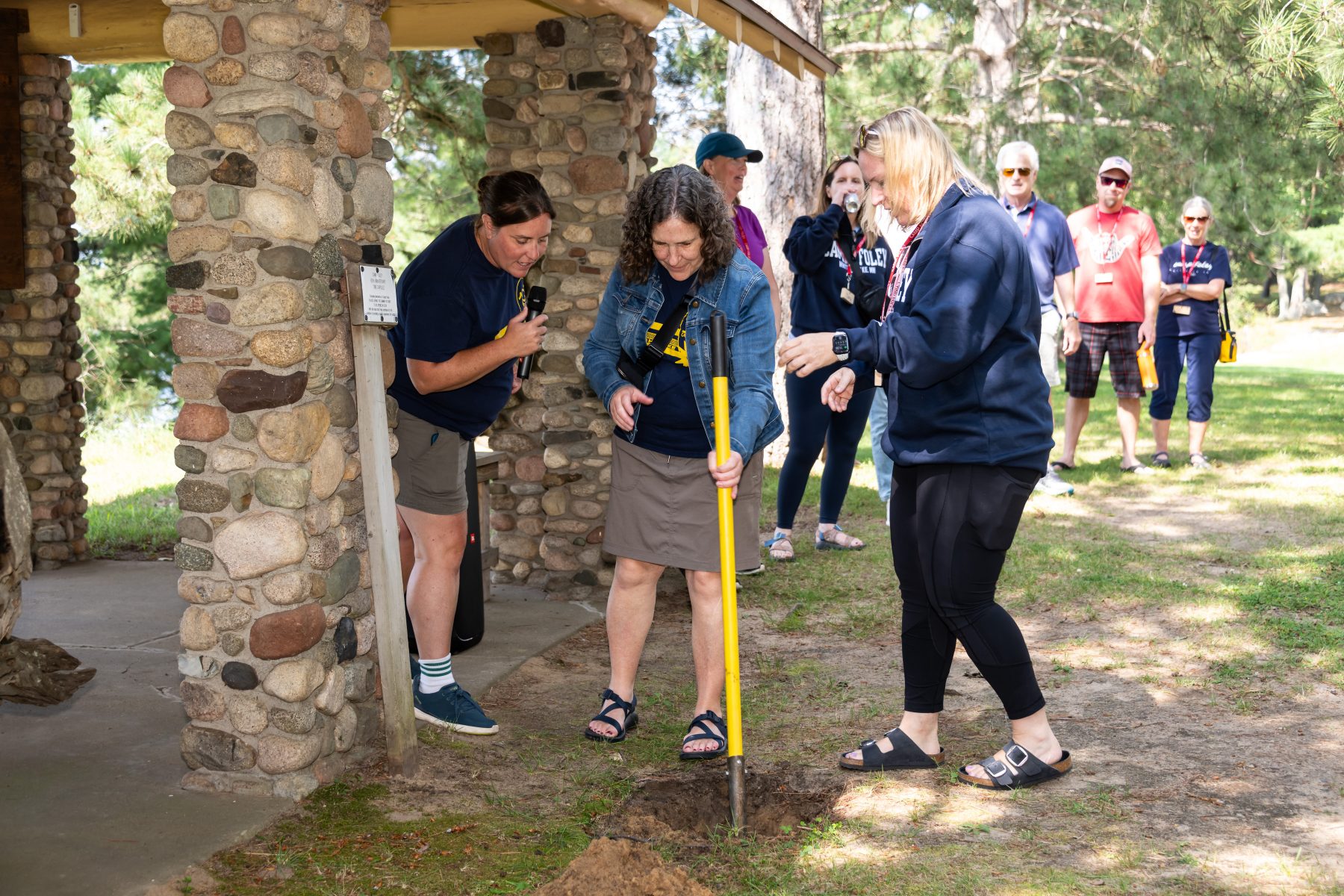 3 women in the center of the photo, the middle woman holds a shovel to start digging up the Camp Foley time capsule