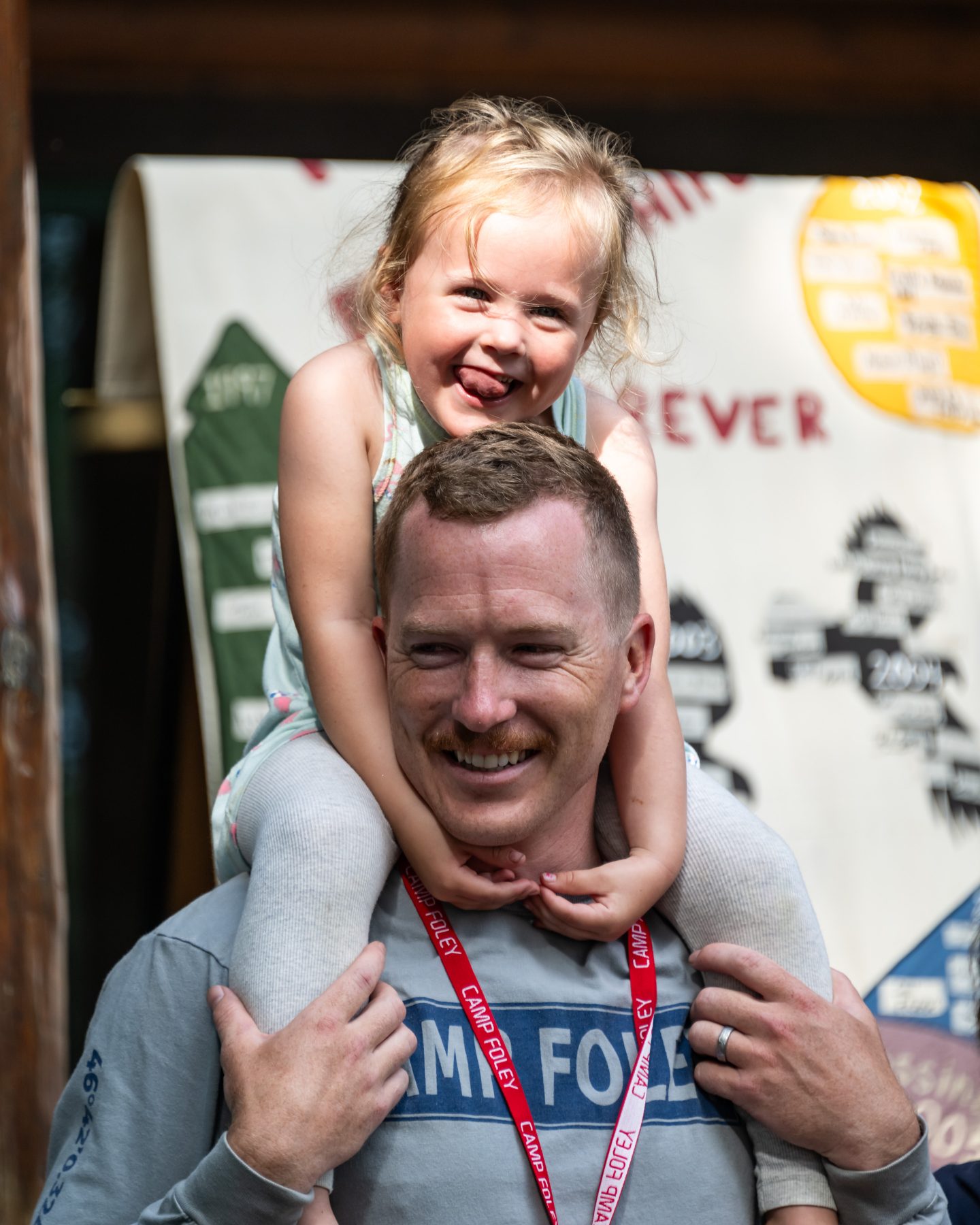 A young girl sits on her dads shoulders, dad is wearing a Camp Foley shirt, both a smiling 