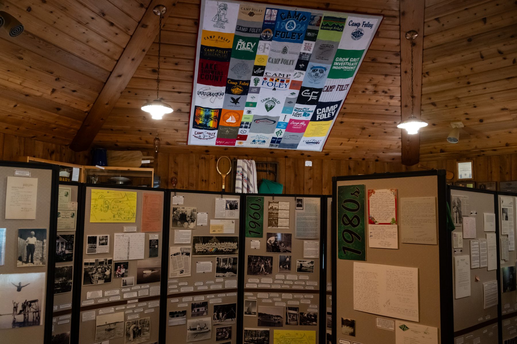 View of a room with wooden panel roof. A quilt hangs from the ceiling and there is museum display boards displaying different decades