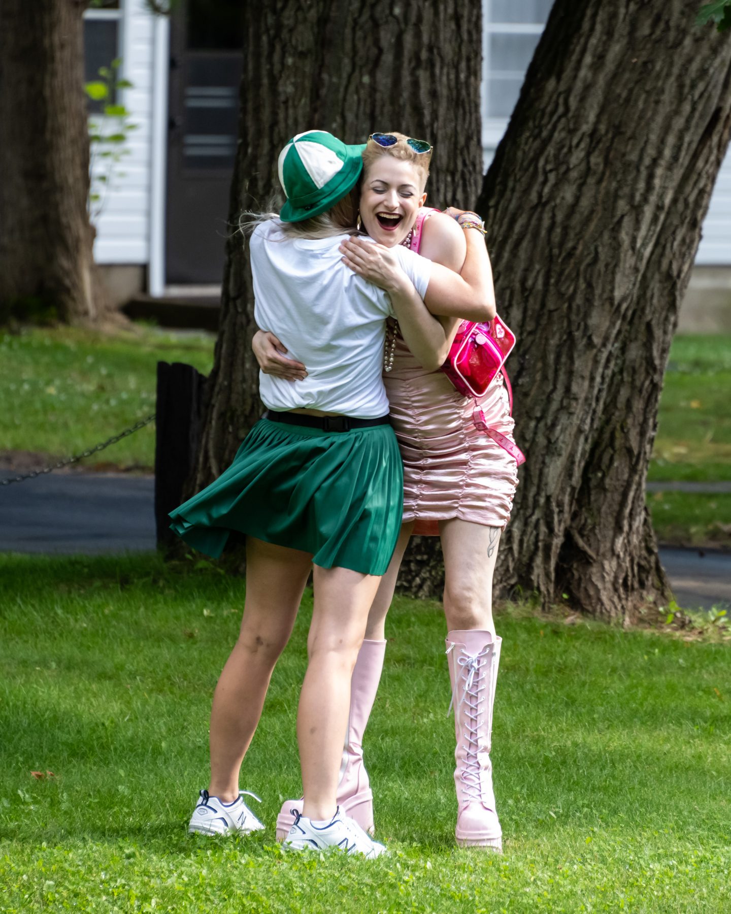 two women are hugging on grass in front of a tree. 