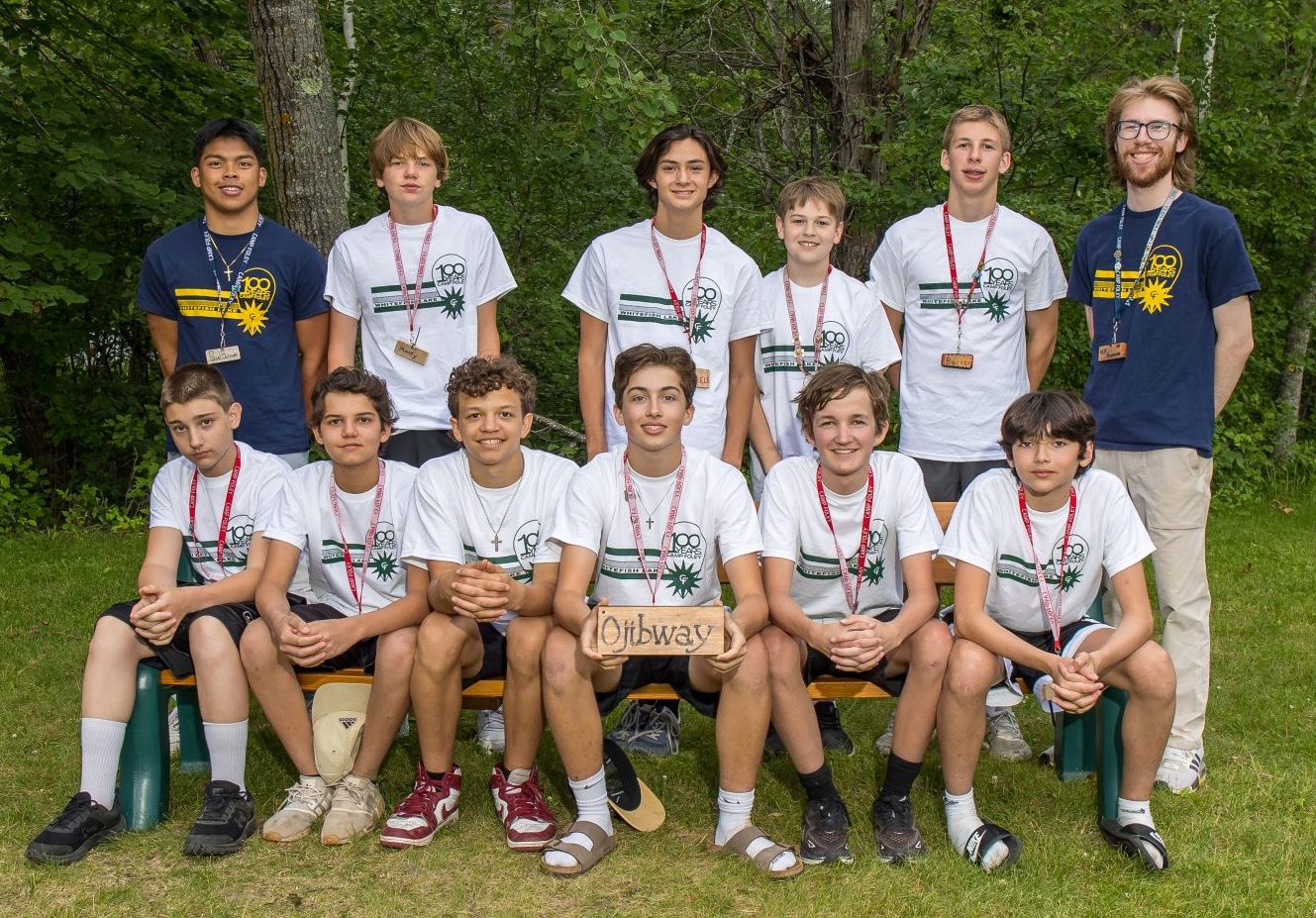 A group of young men sit on a bench all wearing white Camp Foley Shirts, with their counselors standing behind them in navy shirts 