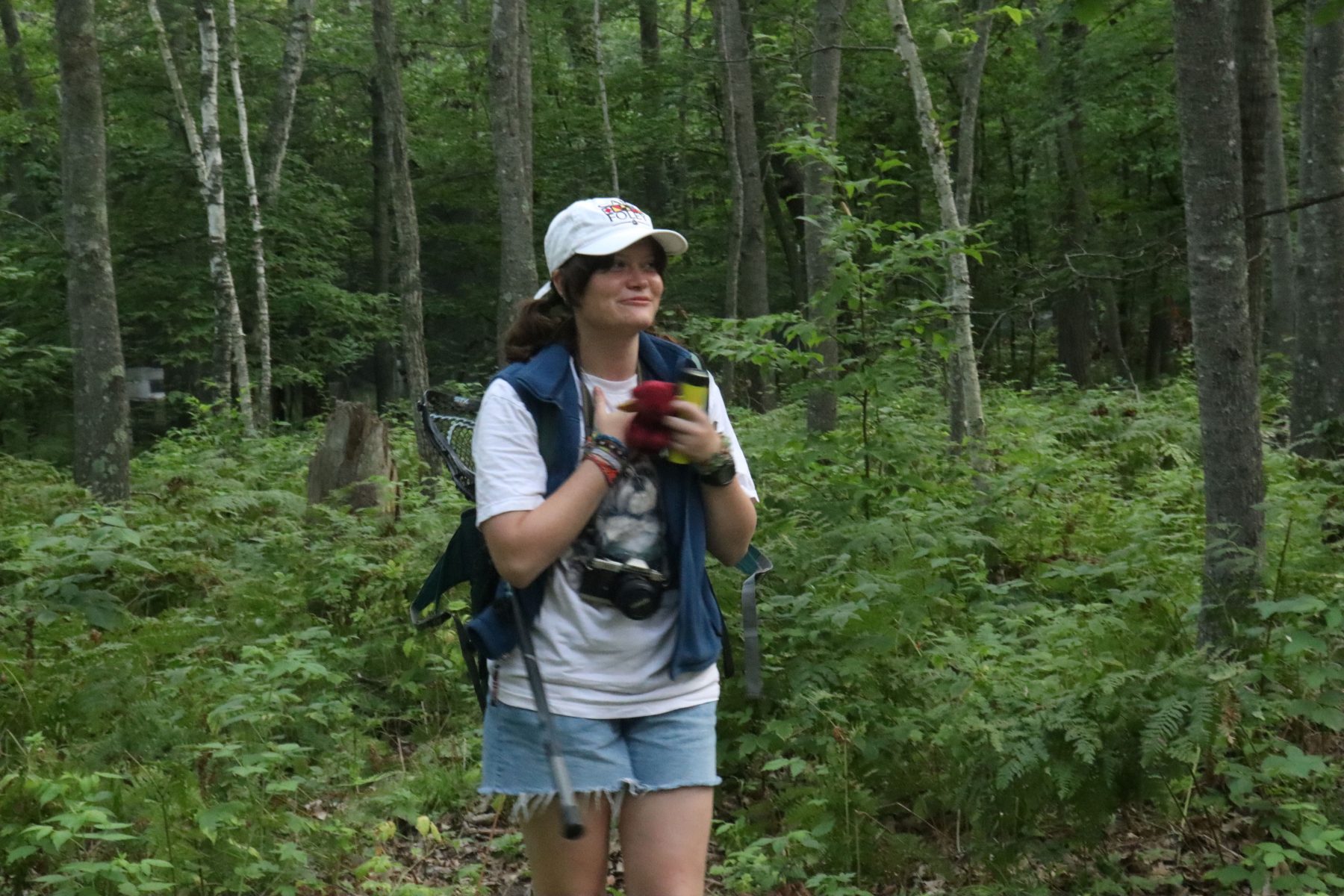 A young woman wearing a denim vest stands in front of trees 