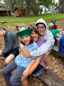 3 brothers sit stacked on each others laps on the Camp Foley benches grinning at the camera 