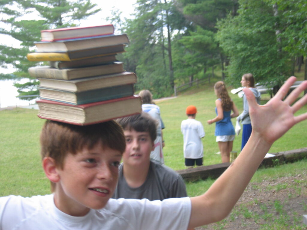 a camp foley boy camper balances several books on his head and gestures off camera 