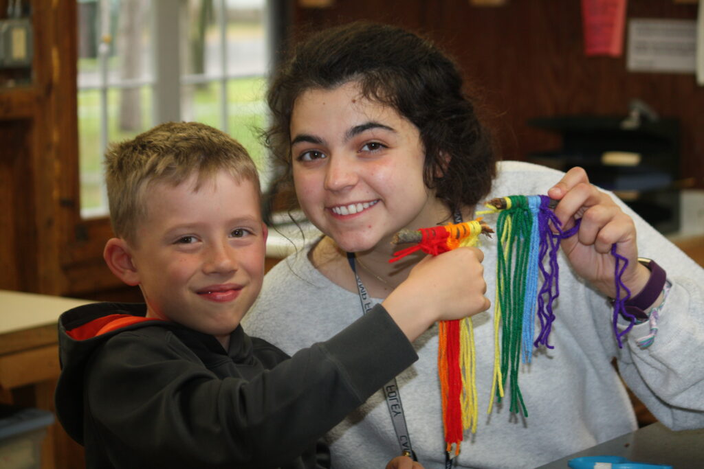 A boy camper and Foley counselor pose together holding a yarn craft at summer camp 