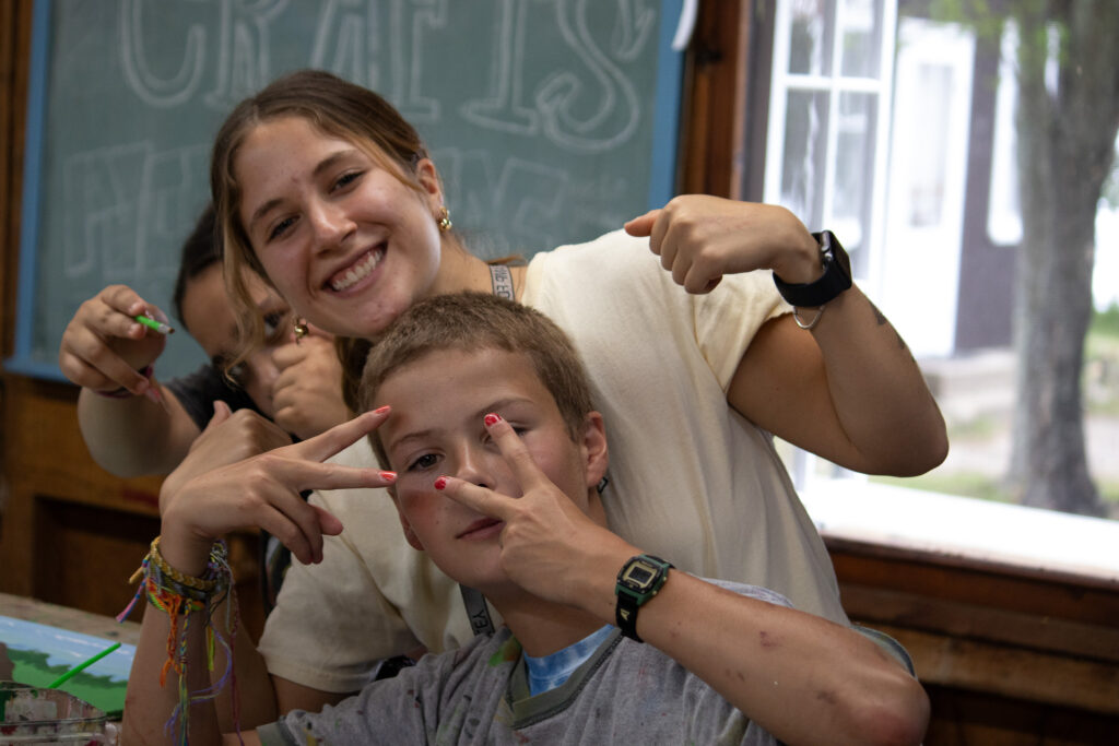 A Camp Foley counselor and camper smile and show peace signs to the camera in the craft building