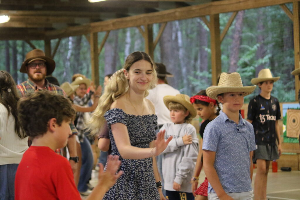 A camp foley counselor square-dances with a camper in a red shirt, with other square dancers around her
