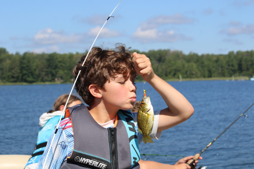 A brunette boy camper leans in to kiss a fish caught on a hook 