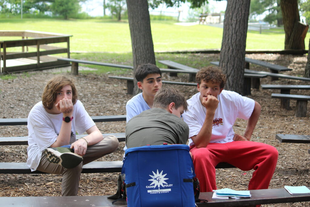 4 boy campers sit on benches, one sits in a Camp Foley camp chair