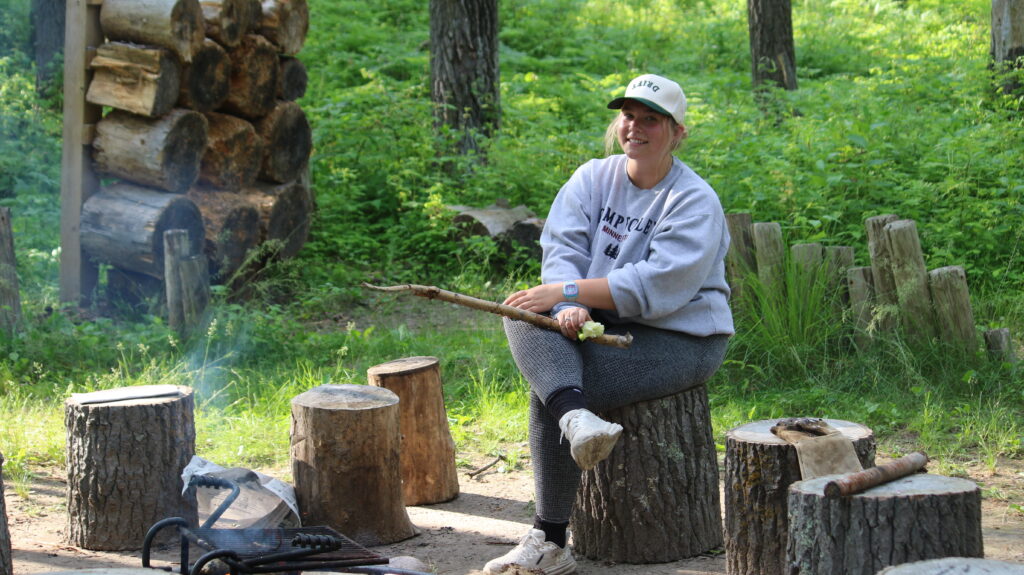 a Camp Foley counselor sits on a stump tending a fire and smiling at the camera